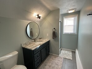A bathroom with a toilet, sink, and mirror. There is a towel rack with a towel hanging on it. The walls are painted in a light color, and there is a window with blinds. - Walk to Beach with with central AC! (Narragansett)