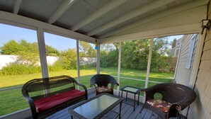 A covered porch with wicker furniture, a glass table, and a view of a green lawn and trees. - Walk to Beach with with central AC! (Narragansett)
