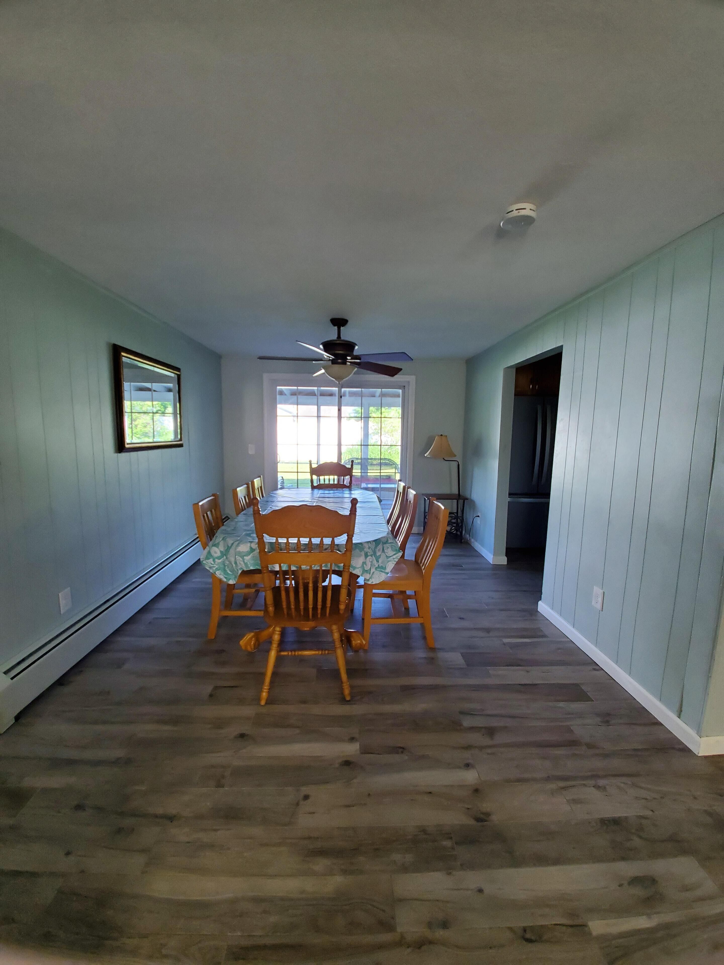 A dining area with a wooden table and chairs, a ceiling fan, and a view through large windows.