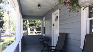 Eine Veranda mit zwei Schaukelstühlen und einer Bank, eine weiße Tür sowie Blick auf einen umzäunten Garten.