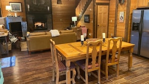 Un interior acogedor de cabaña con una mesa de comedor de madera, sillas, chimenea, zona de cocina y una escalera.