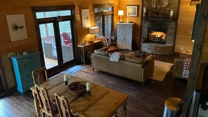 Interior acogedor de una cabaña con comedor, sofá, chimenea y vistas a través de un ventanal.