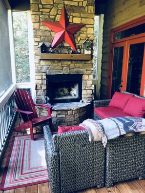 Une cheminée en pierre ornée d’une étoile rouge, un canapé en osier avec des coussins rouges et un fauteuil Adirondack rouge sur une terrasse en bois.