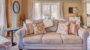 A cozy living room with a beige sofa, patterned cushions, a clock on the wall, and a 'Farmhouse' sign above the windows.