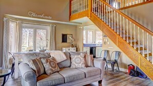 A spacious living room with a large gray sofa, a wooden staircase, and a kitchen in the background.