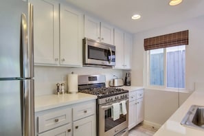 A kitchen with white cabinets, a stainless steel refrigerator, a microwave, a stove, and a window with blinds.