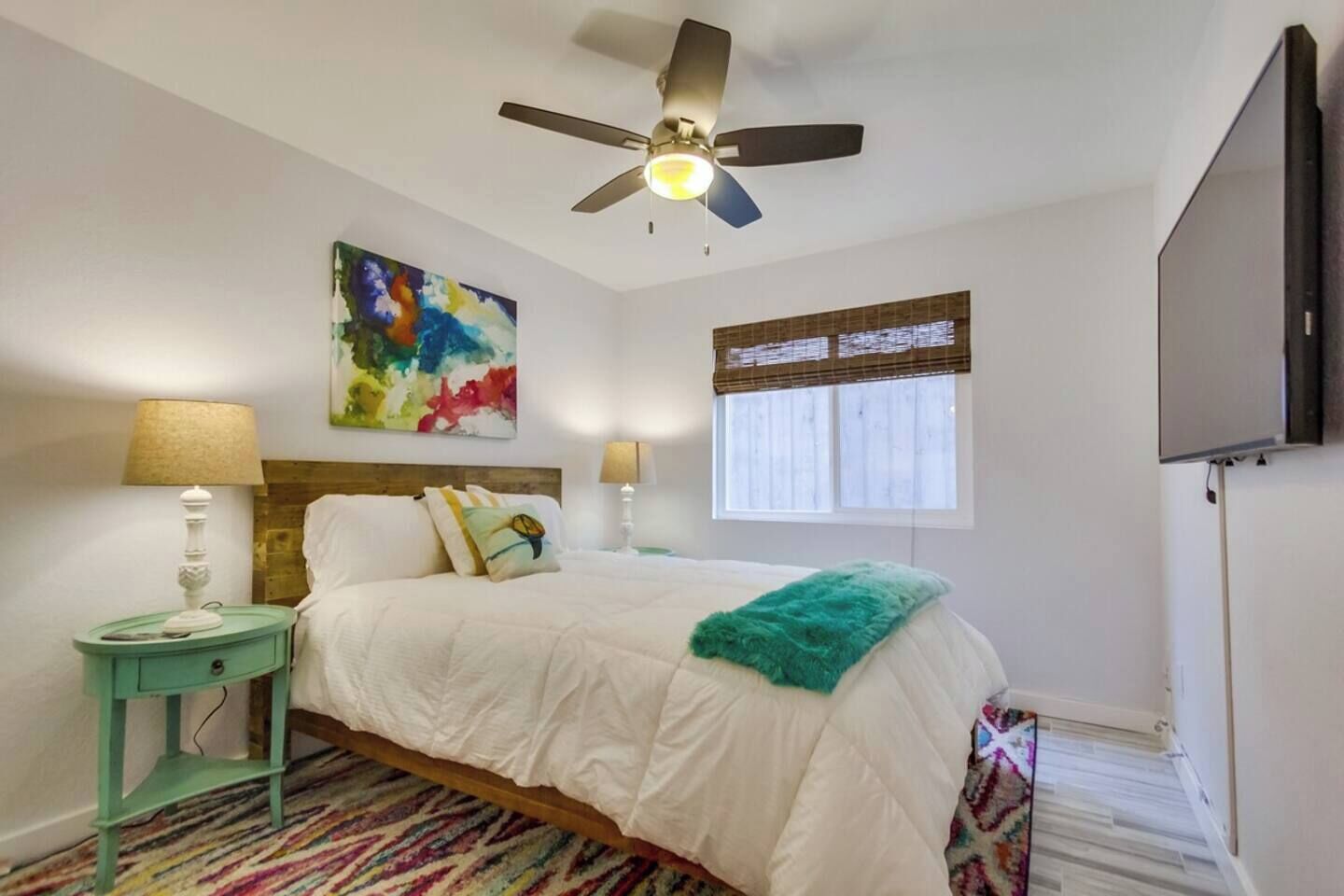A bedroom with a bed, a ceiling fan, a window with blinds, a TV mounted on the wall, and a colorful abstract painting.