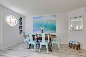 Een eetruimte met een houten tafel, lichtblauwe stoelen, een groot schilderij van een golf aan de muur en een rond raam.