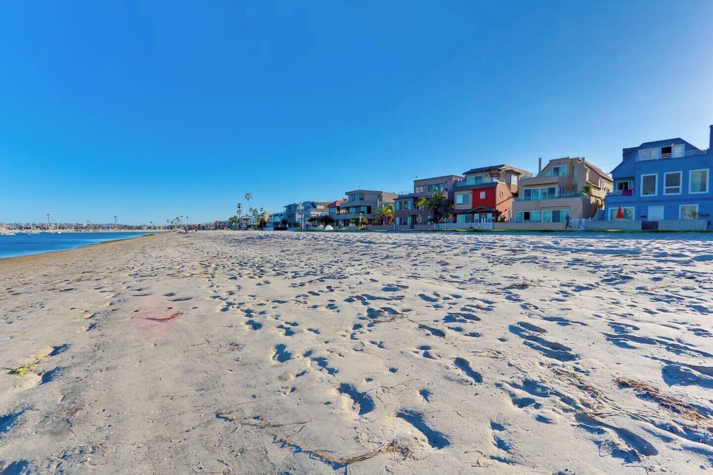 Une plage de sable avec des empreintes, un ciel bleu clair et des bâtiments en arrière-plan.
