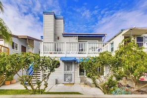 A two-story residential building with a balcony, a garage door, and a small front yard with potted plants.