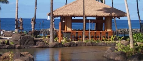 A beachside resort with a thatched-roof hut, lounge chairs, and a pool area surrounded by rocks and palm trees.