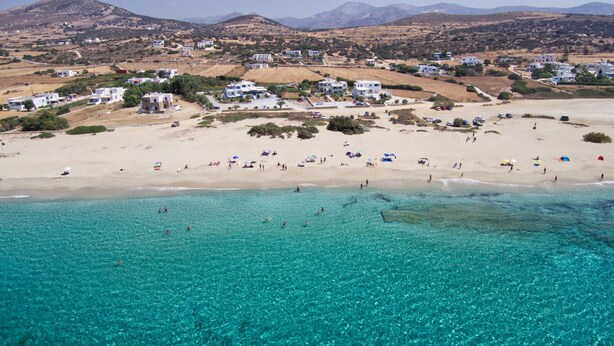 Vista aérea de una playa con aguas turquesas cristalinas, orilla arenosa y edificios blancos al fondo.