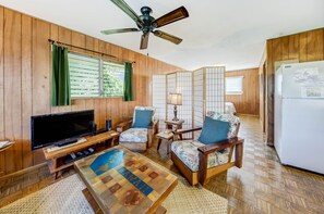 A wooden-paneled room with a television, a coffee table, two armchairs, a ceiling fan, and a refrigerator. - Malolo Cottage - a studio/1ba home near the beach in Hanalei (Hanalei)