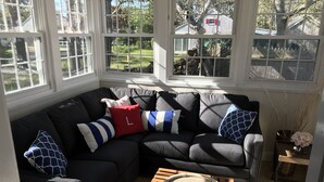 A cozy living room with a dark-colored sectional sofa, multiple pillows, a small wooden table with a mirror, and a view of a garden through large windows.