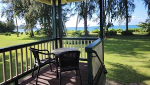 A covered outdoor seating area with a table and two chairs, overlooking a grassy landscape with trees and a view of the ocean. - Ka Nani, steps from the sand! The center of Hanalei Bay. Best $ on the bay. (Hanalei)