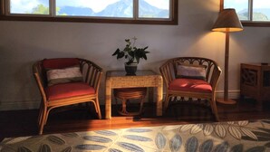 A room with two wicker chairs, a small table with a potted plant, a floor lamp, and a view of mountains through large windows. - Ka Nani, steps from the sand! The center of Hanalei Bay. Best $ on the bay. (Hanalei)