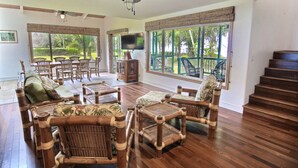 A spacious living room with a wooden floor, a ceiling fan, and large windows offering a view of the outdoors. - Ka Nani, steps from the sand! The center of Hanalei Bay. Best $ on the bay. (Hanalei)