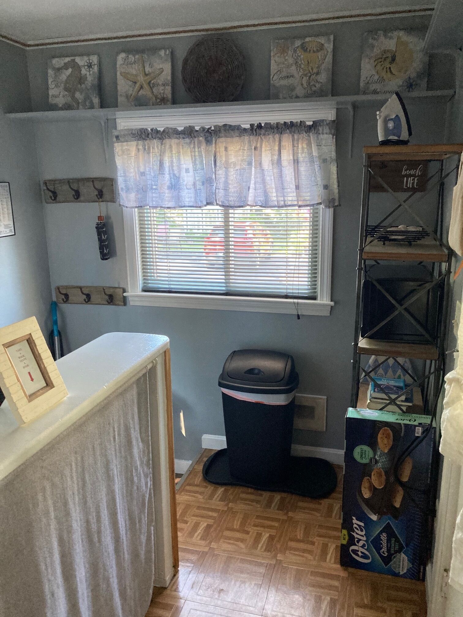 A compact kitchen with a window, a sink, a trash bin, and a shelf with kitchen items.
