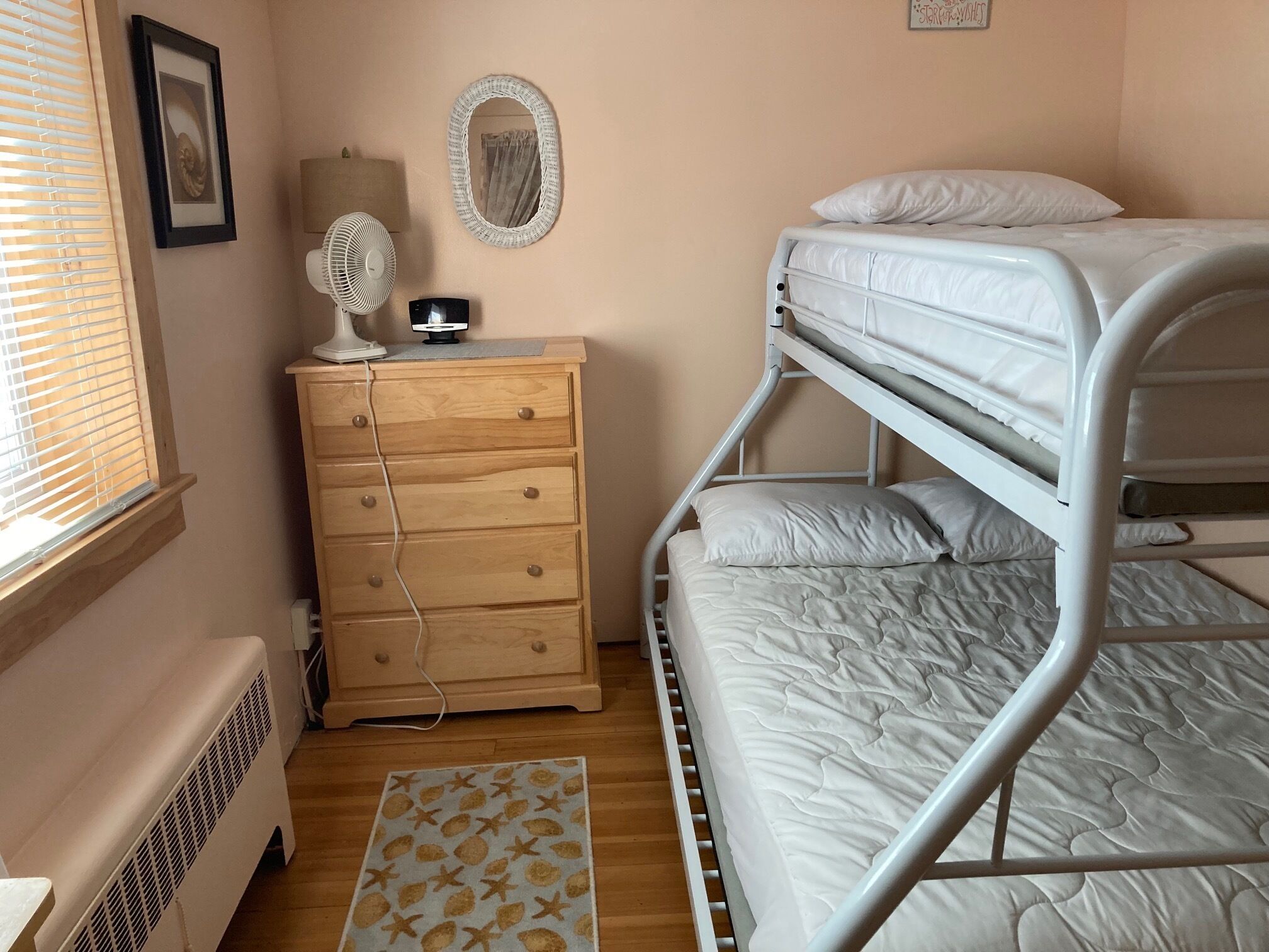 A bunk bed with white bedding, a wooden dresser with drawers, a standing fan, and a small rug with a starfish pattern.