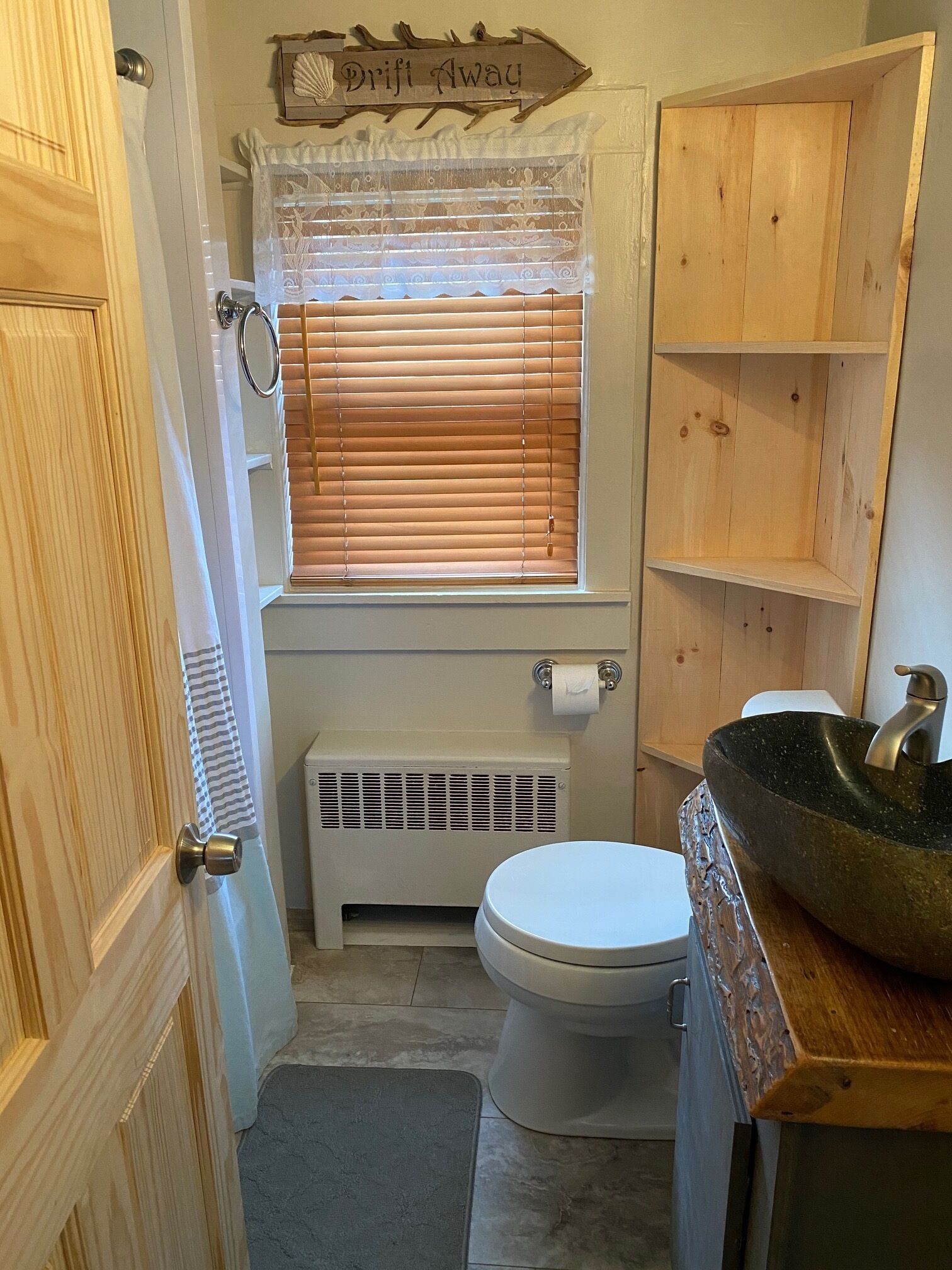 Une petite salle de bain comprenant des toilettes, une fenêtre avec stores, un radiateur et une étagère en bois.