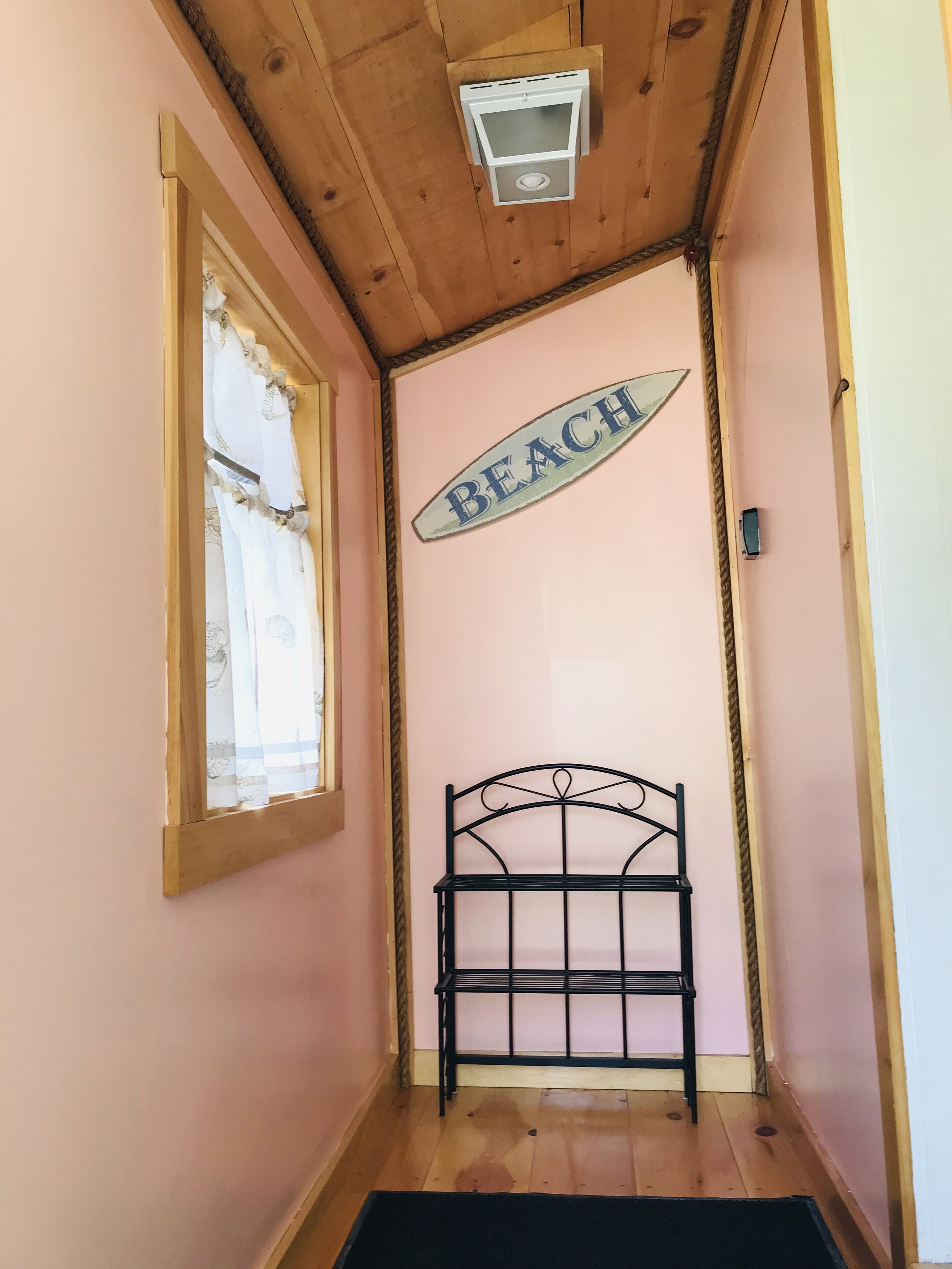 Un couloir étroit avec un plafond en bois, un cadre de lit en métal noir, une fenêtre avec un rideau blanc et un panneau indiquant « PLAGE » sur le mur.