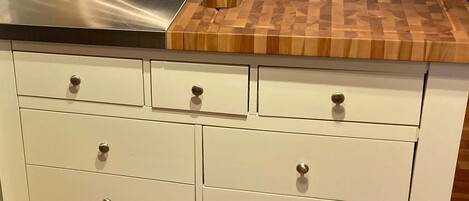 Cocina con campana extractora de acero inoxidable, tabla de cortar de madera y alacenas blancas con varios cajones.
