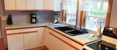 A compact kitchen with white cabinets, a coffee maker, a blender, a sink, and a window with curtains.