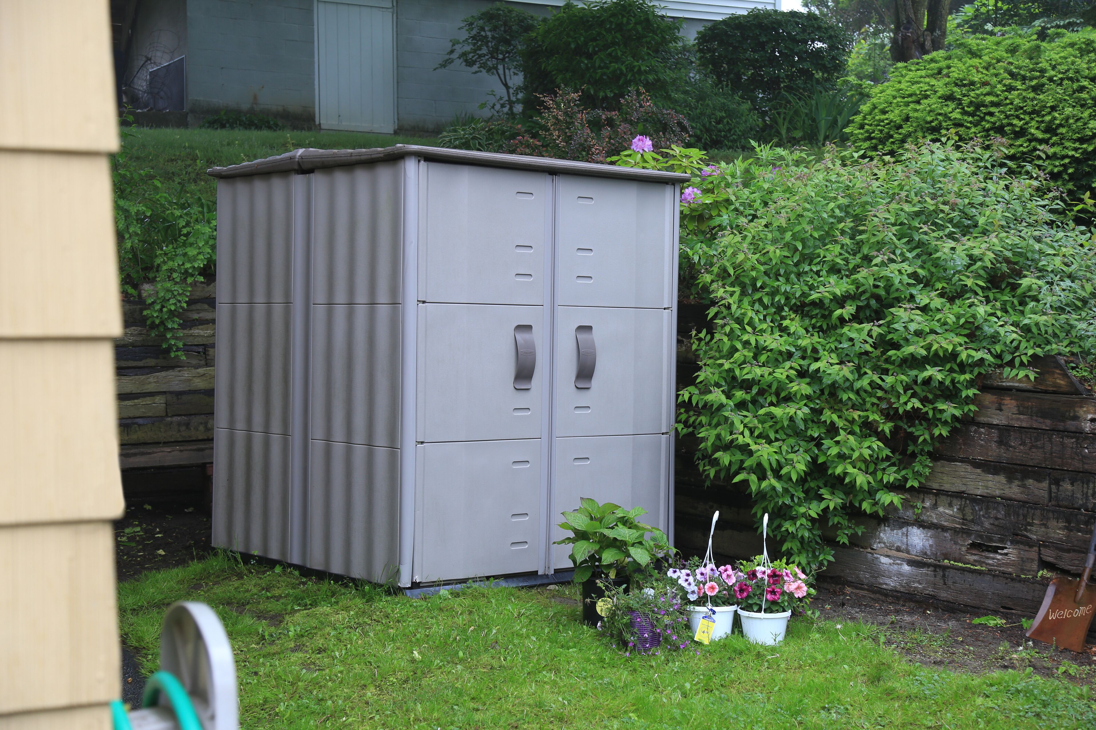 Een grijze tuinhuisje met meerdere deuren en handgrepen, omgeven door een gazon en potplanten.