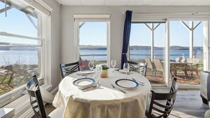 A dining area with a table set for four, large windows offering a view of the sea, and a casual outdoor seating area visible through the windows.