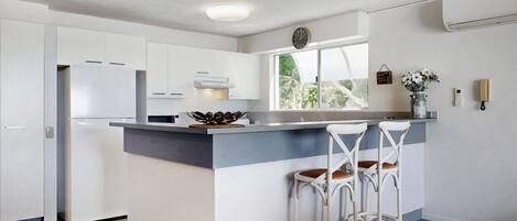 A modern kitchen with white cabinets, a grey countertop, and a window with a view of greenery. There are two white bar stools with brown cushions.
