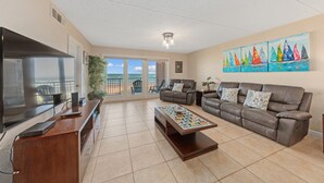 A spacious living room with a large flat-screen TV, a brown leather sofa, a coffee table, and a painting of sailboats on the wall. - SPI Beach House Gateaway 1-504 (South Padre Island)