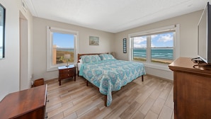 A bedroom with a large bed, two windows with ocean views, a wooden dresser, and a nightstand. - SPI Beach House Gateaway 1-504 (South Padre Island)