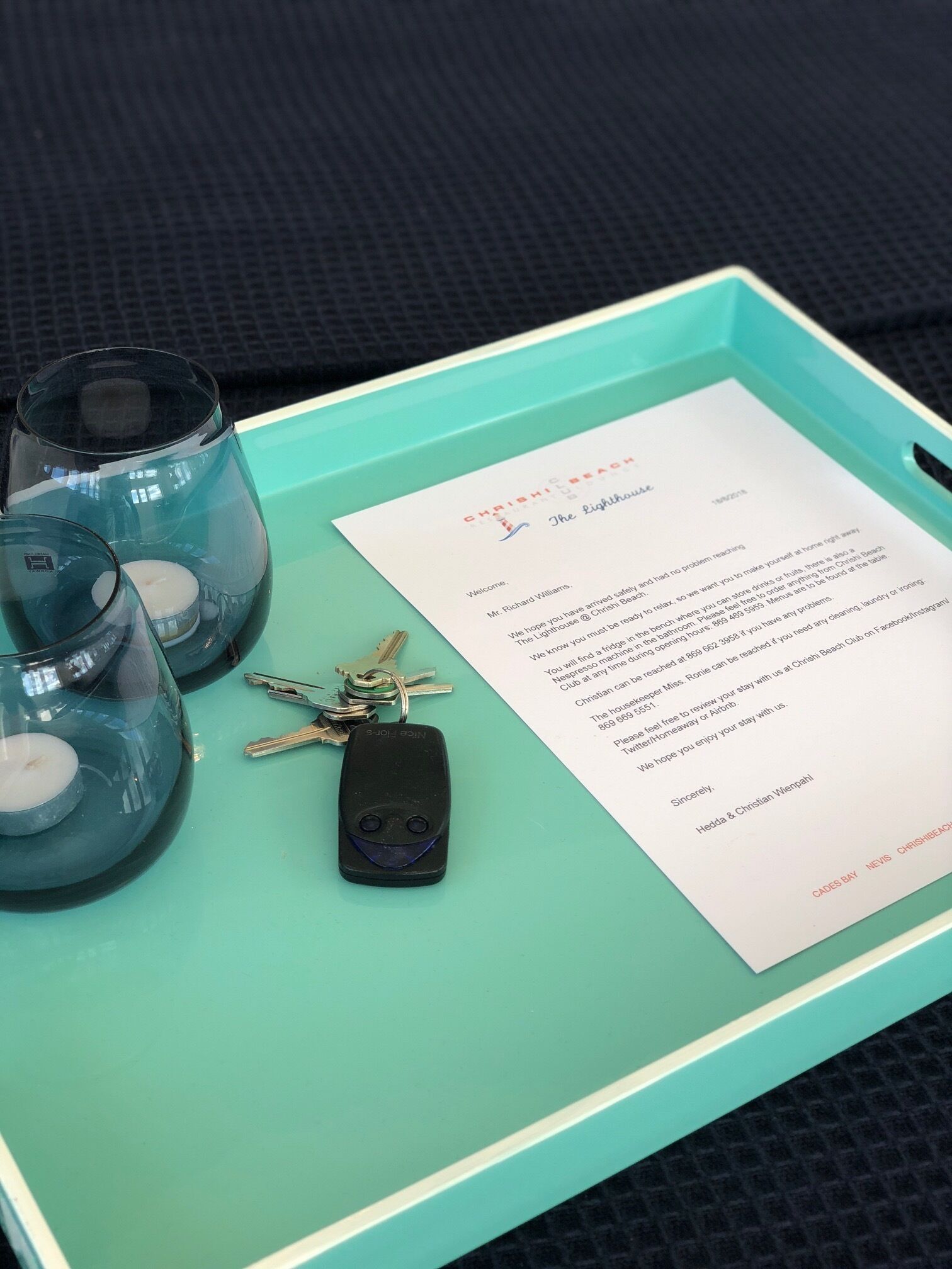 A teal tray with three glass candle holders, a set of keys, and a welcome letter from a hotel.