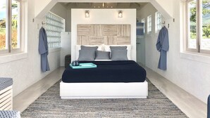 A modern bedroom with a large bed, a headboard with a wood panel design, and a view of the outdoors through large windows. - Unique Beachfront Suite, private with an amazing view w option of 2 more rooms. (Cades Bay)