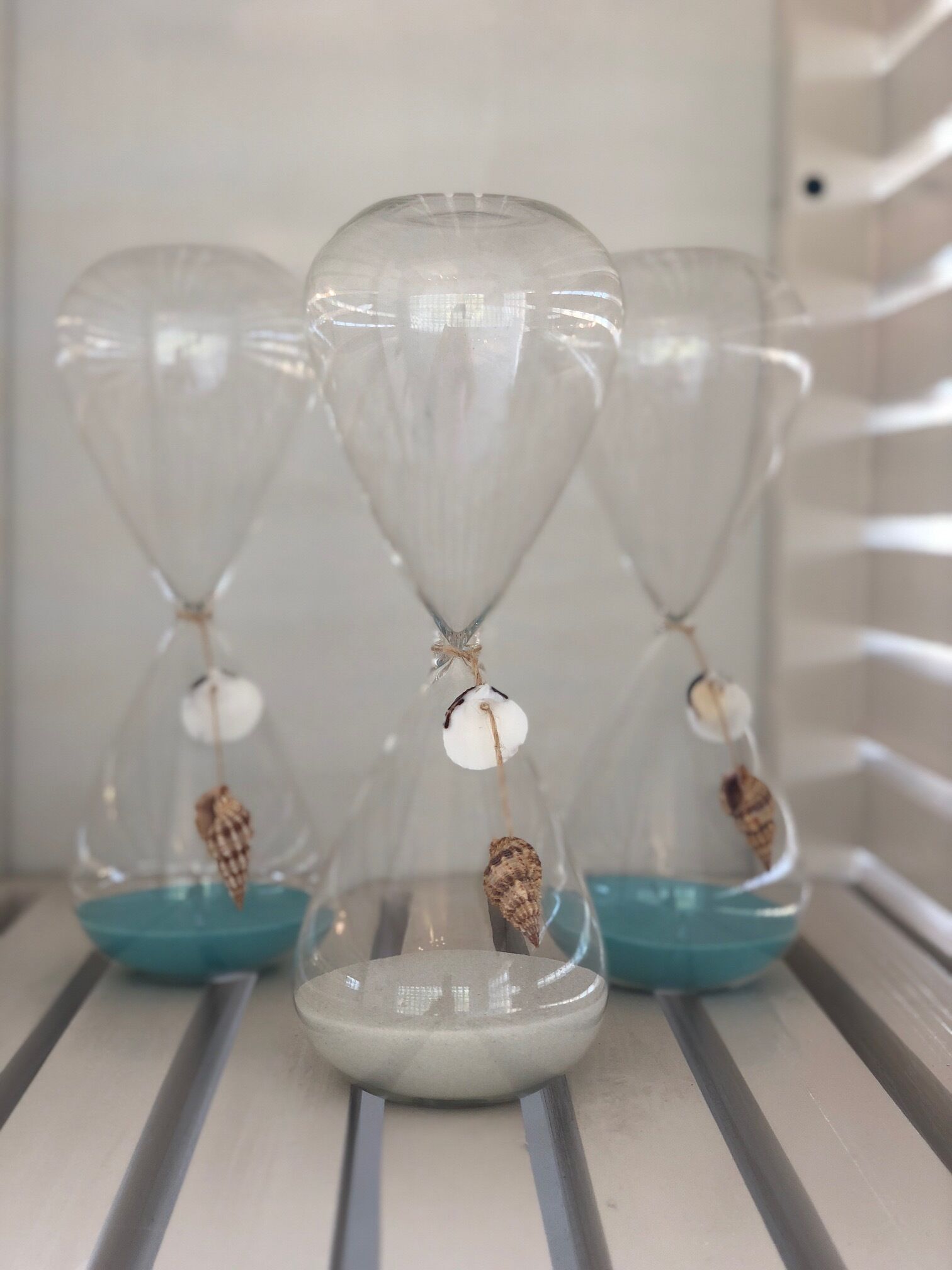Tres relojes de arena con conchas y una base blanca sobre una estantería.