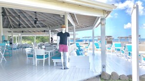 A covered outdoor seating area with white tables and chairs, a mannequin, and a view of the beach. - Unique Beachfront Suite, private with an amazing view w option of 2 more rooms. (Cades Bay)