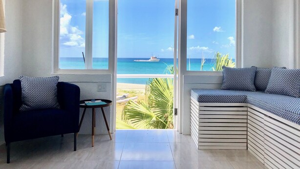 A room with a large sliding glass door leading to a balcony with a view of the ocean and a boat. There is a dark blue sofa with patterned cushions and a small round table with a green top. - Unique Beachfront Suite, private with an amazing view w option of 2 more rooms. (Cades Bay)