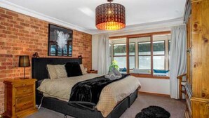 A bedroom with a brick wall, a bed with a black headboard, a wooden nightstand, a lamp, a window with curtains, and a chandelier. - Hat Hill Hideaway Amazing Valley Views (BLACKHEATH)