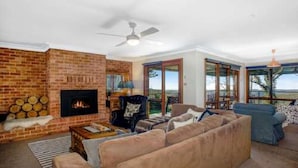 A living room with a brick fireplace, a ceiling fan, and a large window offering a view of the outdoors. - Hat Hill Hideaway Amazing Valley Views (BLACKHEATH)