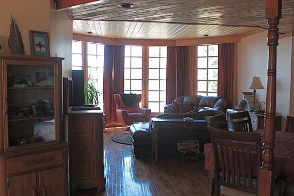 Amplia sala de estar con piso de madera, un sofá de cuero, un sillón rojo, una mesa de comedor con sillas y un ventanal con cortinas.