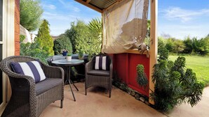 Un patio couvert, aménagé avec des meubles en osier et une table, entouré de verdure et offrant une vue sur une pelouse.
