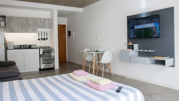 A modern hotel room with a kitchen, a dining area, and a TV mounted on the wall.