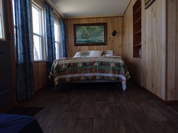 Een kamer met houten wanden, een bed met bloemenbedekking, een schilderij van een schip aan de muur, en een raam met gordijnen.