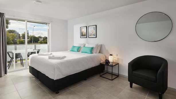 A modern bedroom with a large bed, a black armchair, a round mirror, and two framed pictures on the wall. - East Maitland Executive Apartments (East Maitland)
