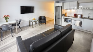 A modern kitchen with white cabinets, a stainless steel refrigerator, and a microwave. There is a dining area with a table and chairs, and a black leather sofa. - East Maitland Executive Apartments (East Maitland)