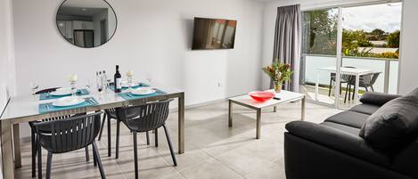 A modern living room with a dining area, a black leather sofa, a glass table, and a balcony with a view.