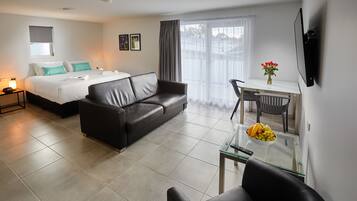 Quarto de hotel moderno com cama, sofá de couro, mesa de centro de vidro, mesa de jantar com cadeiras e televisão na parede.