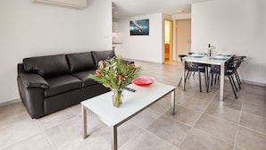 A modern living room with a black leather sofa, a white coffee table with a vase of flowers, and a dining area with a table set for two. - East Maitland Executive Apartments (East Maitland)