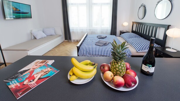 A hotel room with a bed, a sofa, a dining table with fruit and magazines, and a bottle of wine. - Black and White , Wifi and Parking free (Praha 3)