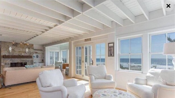 A spacious living room with a high ceiling, a fireplace, and large windows offering a view of the ocean. - Beachfront Property on Private Moody Beach (Wells)